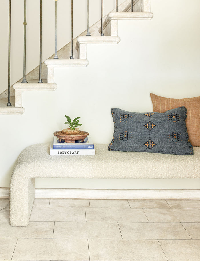 #color::blue #color::indigo #insert::down #insert::polyester | The Indigo woven cotton boho and folk-inspired lumbar pillow with a wool front sits on a white boucle bench with a stack of books and an orange throw pillow