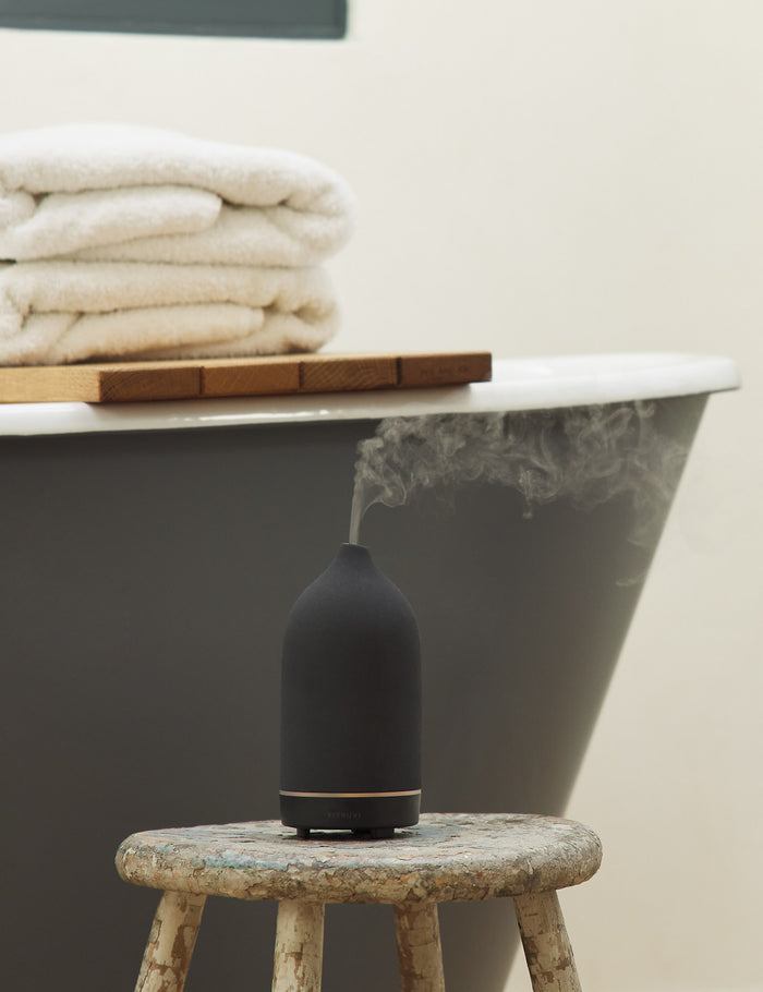 #color::black | The Black Stone Diffuser by Vitruvi sits atop a wooden stool in a bathroom with a black painted tub