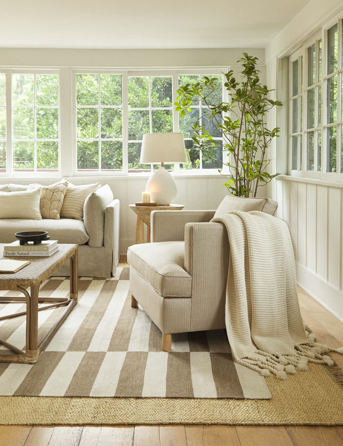 #color::white | The Trestles white chunky knit throw by pom pom at home lays atop a sand-toned accent chair in a living room with a woven coffee table and a brown and cream striped rug
